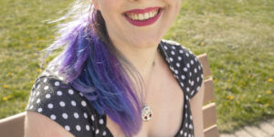 Lynne sits on a wooden bench surrounded by green. They have blue and purple hair, are smiling, and wear a black and white polkadotted dress.